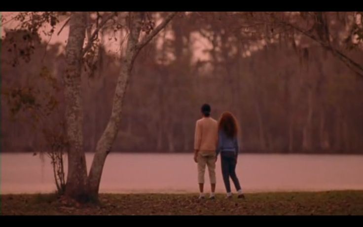 two people standing next to each other in front of a lake and trees with water behind them