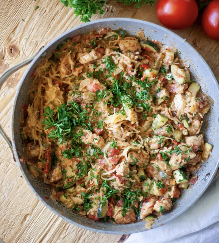 a skillet filled with pasta and vegetables on top of a wooden table next to tomatoes