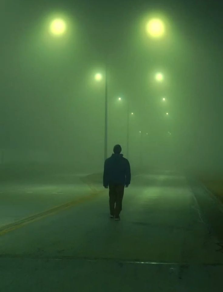 a person walking down a street at night in the fog with three lights on either side of him