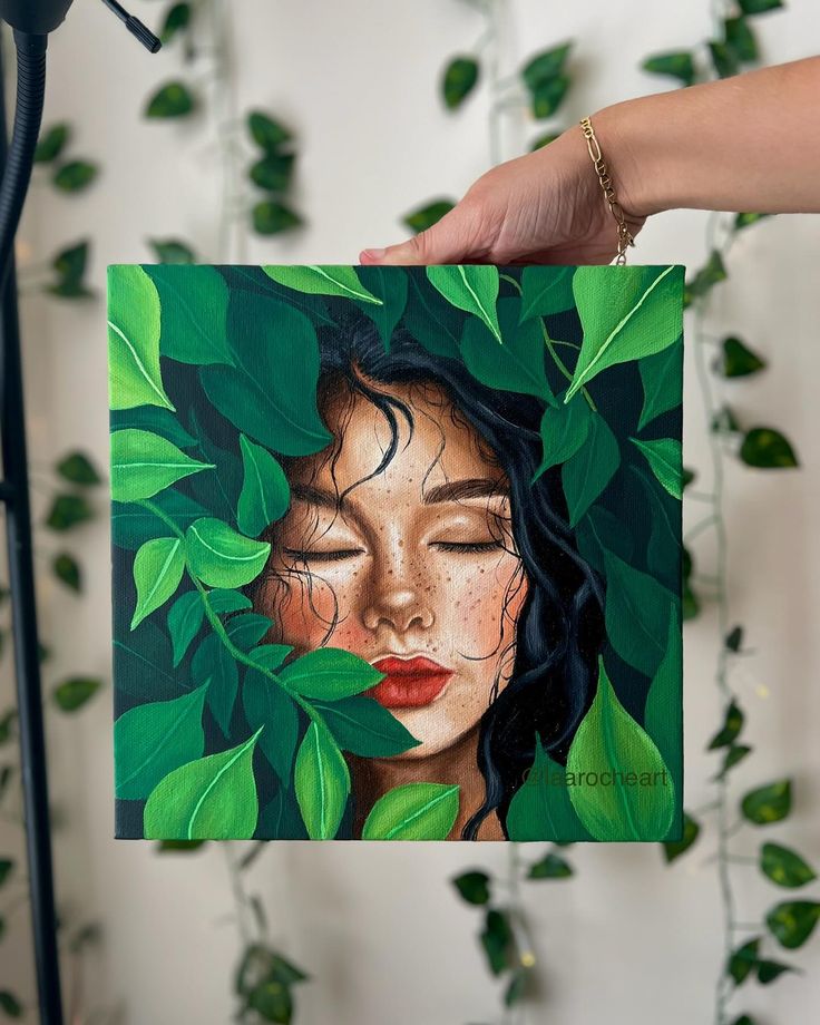 a woman's face is surrounded by green leaves as she holds up a piece of art