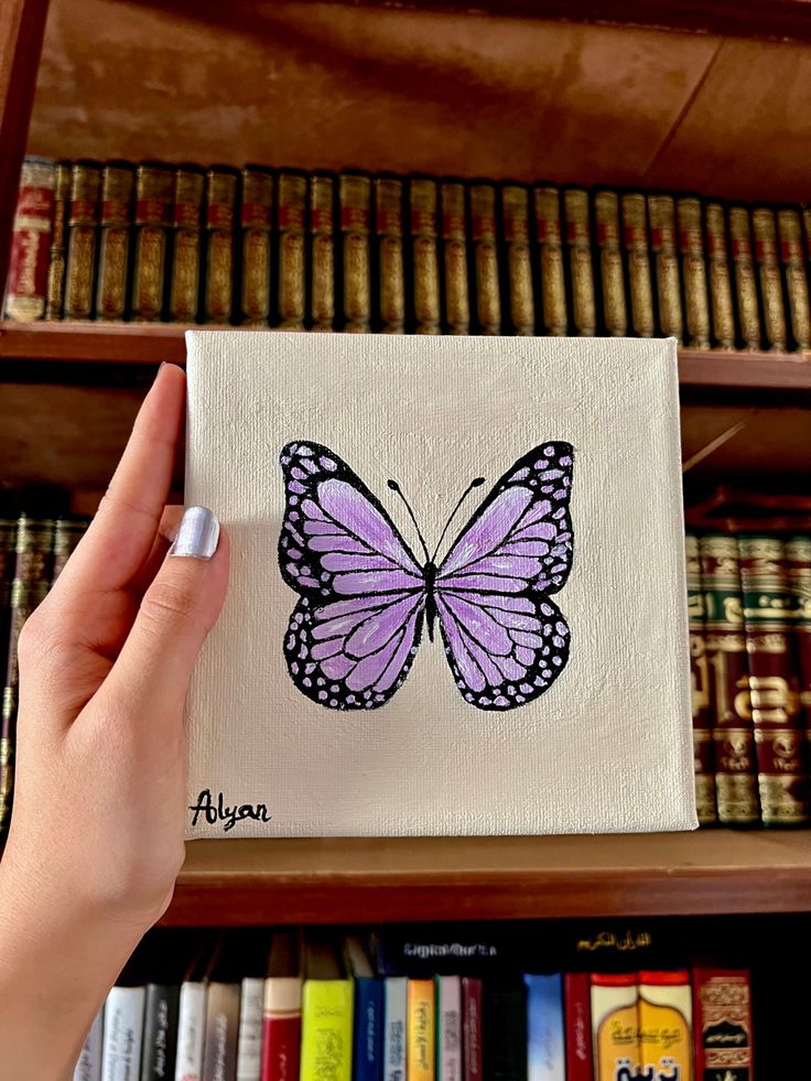 a hand is holding up a small purple butterfly on a white canvas in front of bookshelves