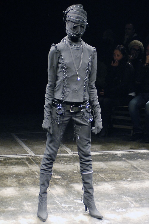 a man in grey is standing on the runway with his head covered by chains and boots