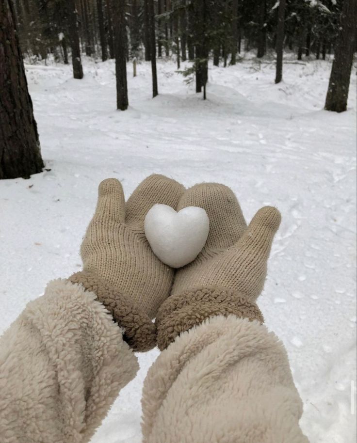 a person wearing gloves holding a white heart in the middle of a snowy forest area