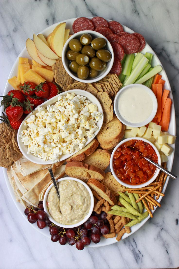 a platter filled with cheese, crackers, fruit and dips on a marble table