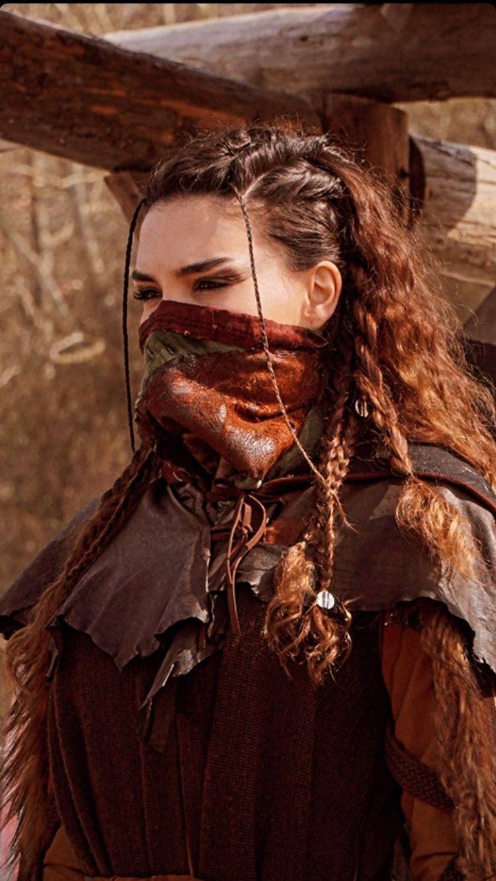 a woman with long hair wearing a brown outfit and braids on her face is standing in front of a wooden structure