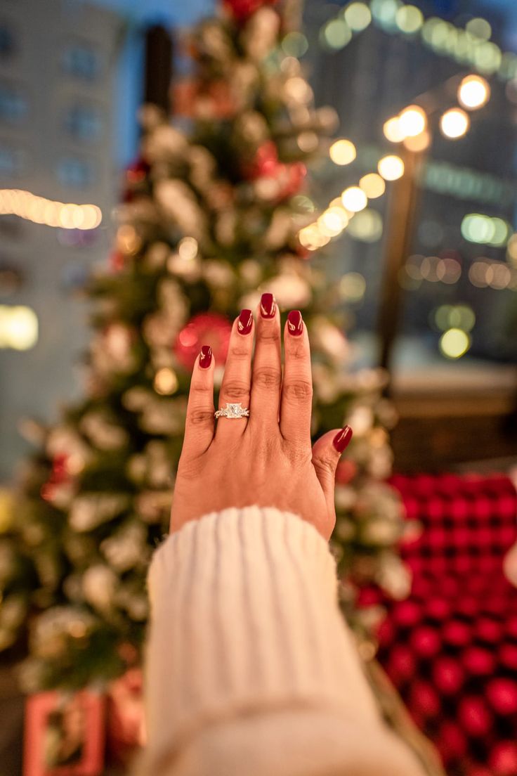 a woman's hand with red nail polish holding a diamond ring in front of a christmas tree