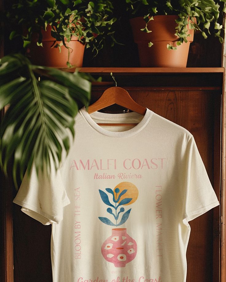 a white t - shirt with an image of a pink vase on the front and green plants behind it