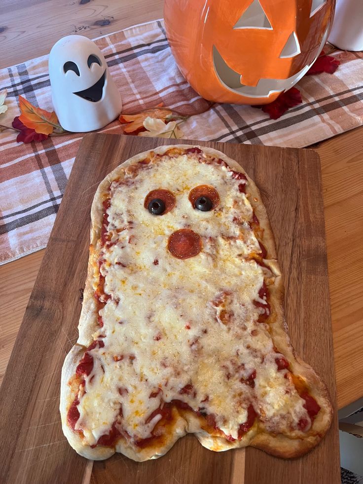 a pizza shaped like a ghost sitting on top of a wooden cutting board next to a pumpkin