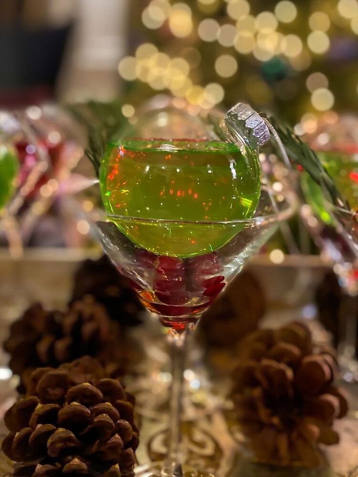 two glasses filled with green liquid next to pine cones and christmas lights in the background