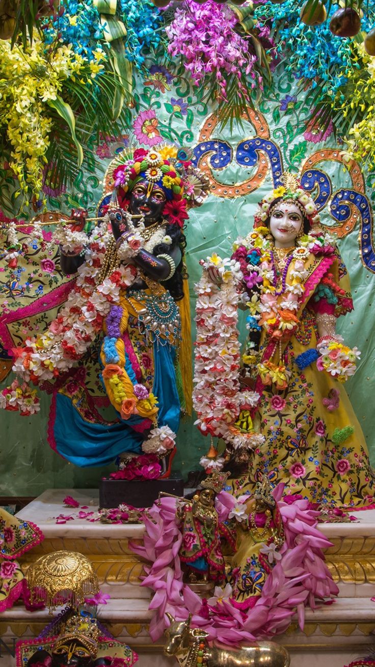 two deities dressed in colorful clothing standing next to each other with flowers on the wall behind them