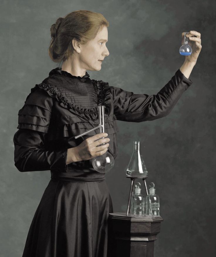 a woman in black dress holding a glass beaker and flasks on top of a table