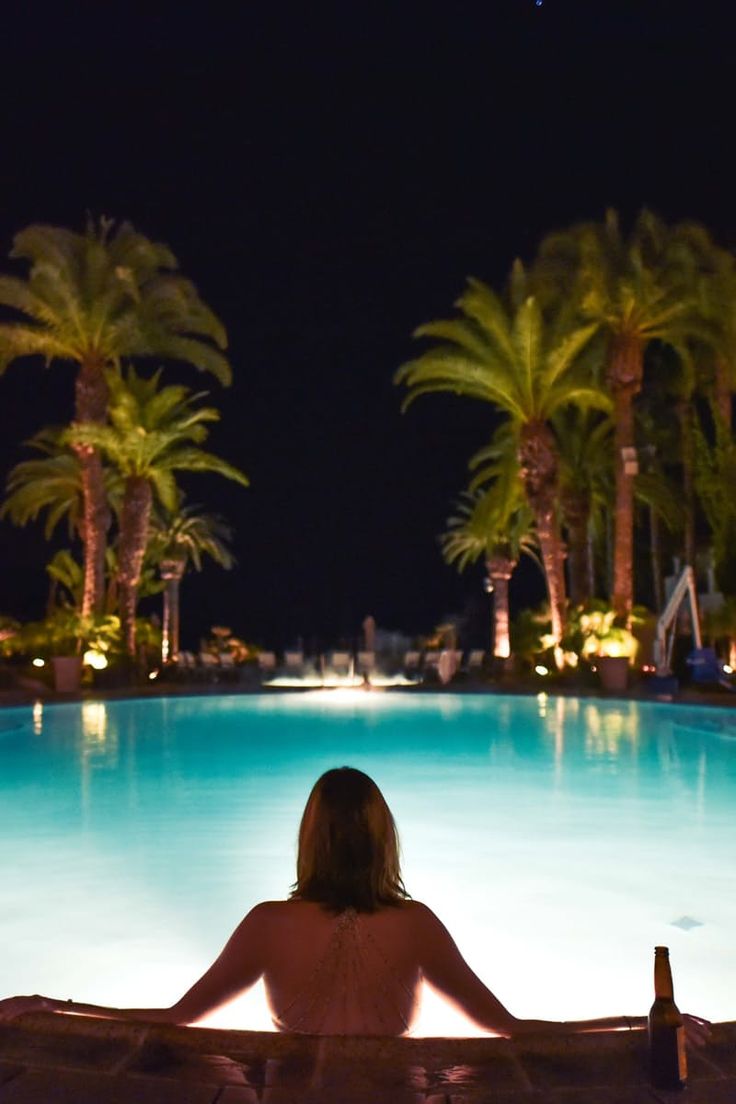 a woman sitting on top of a wooden bench next to a swimming pool at night