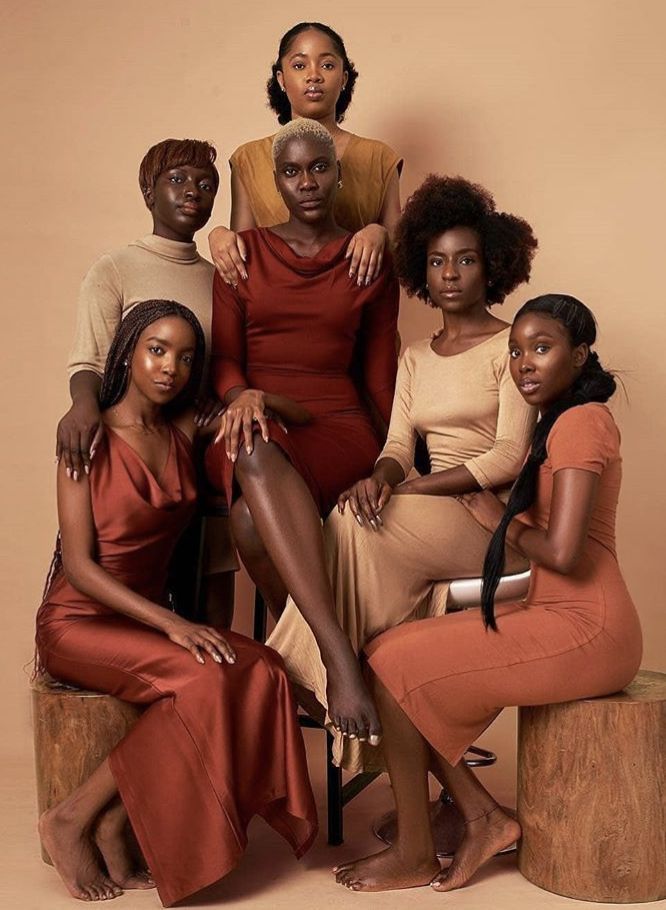 four women sitting on stools in front of a beige background, one is wearing a red dress and the other wears an orange top