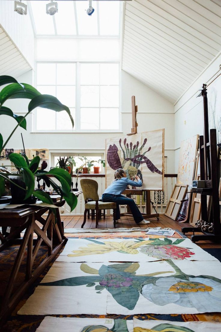 a person sitting at a table in an art studio with paintings on the wall and floor