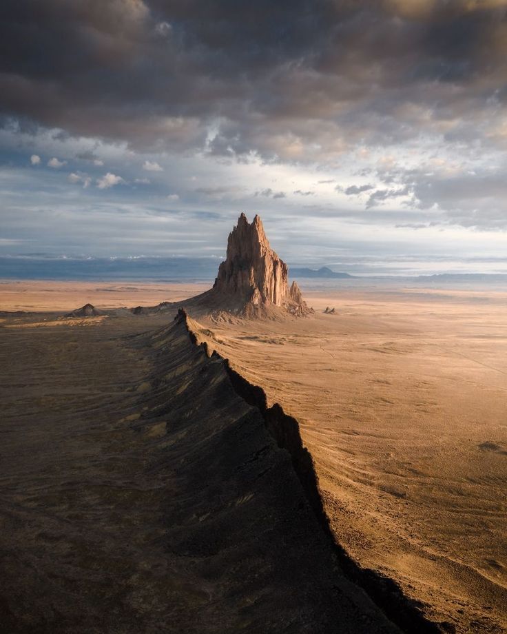 a large mountain in the middle of a desert under a cloudy sky with dark clouds