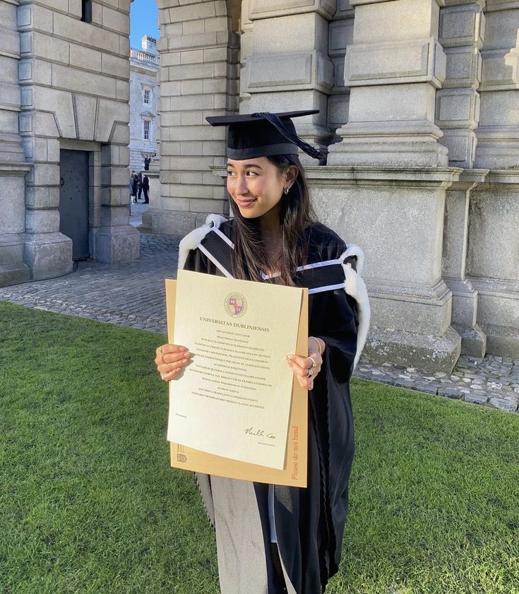 a woman in graduation gown holding up her diploma and standing on the grass outside an old building