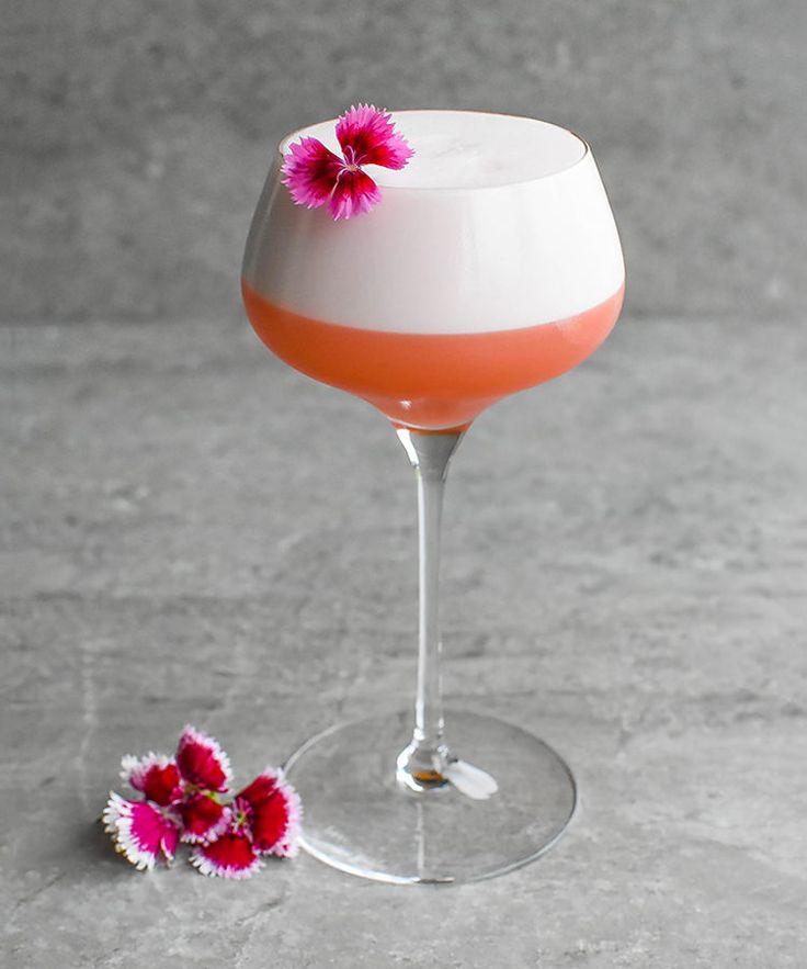 an orange and white drink in a wine glass with pink flowers on the rim next to it