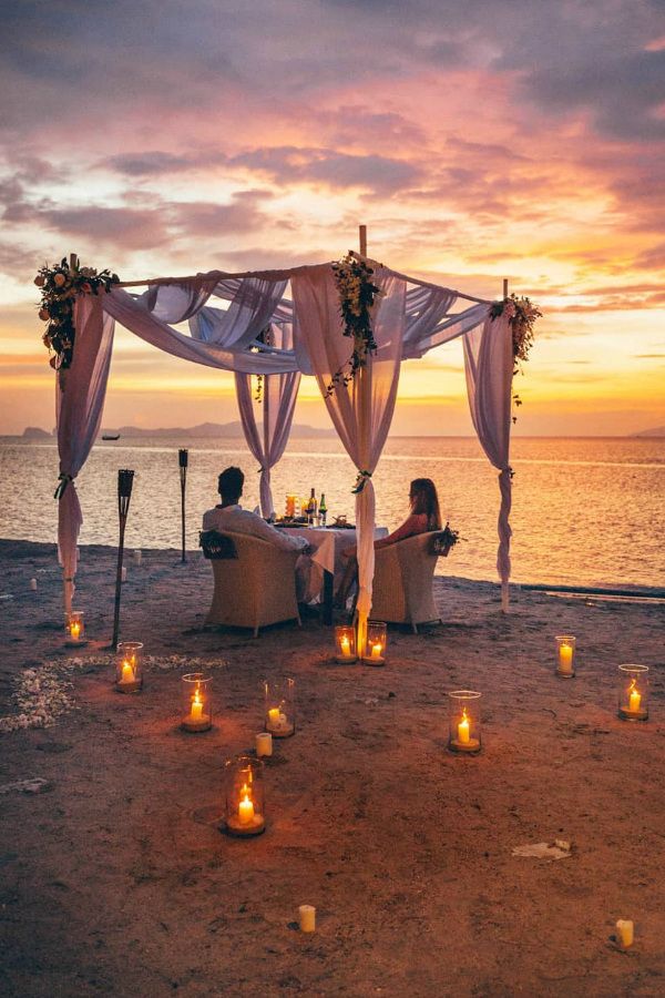 two people sitting at a table with candles in front of them on the beach during sunset