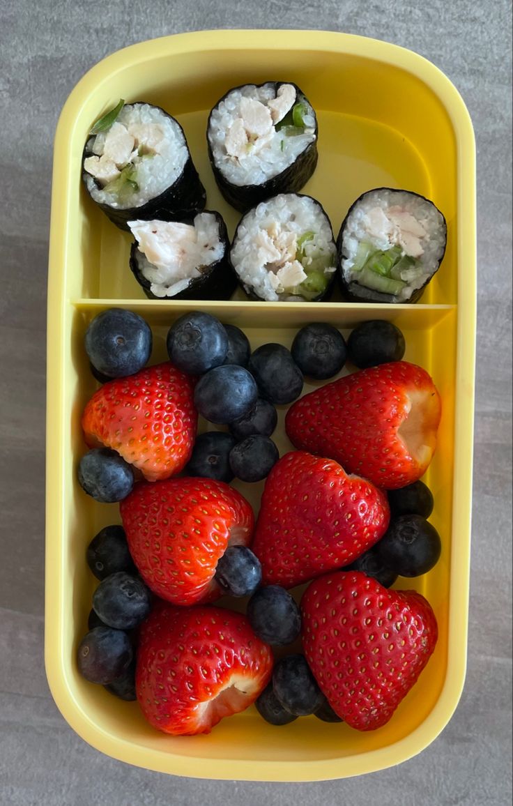 a yellow tray with strawberries, blueberries and sushi in it on a table