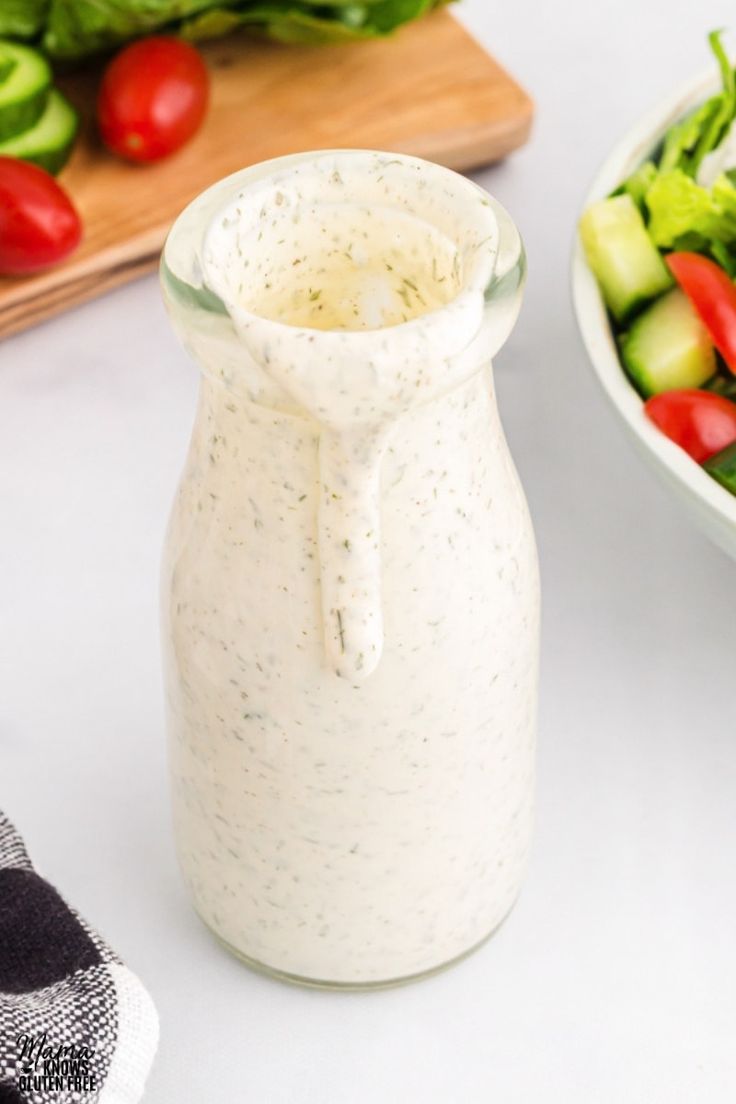 a bowl of salad next to a bottle of dressing on a table with tomatoes, cucumbers and lettuce