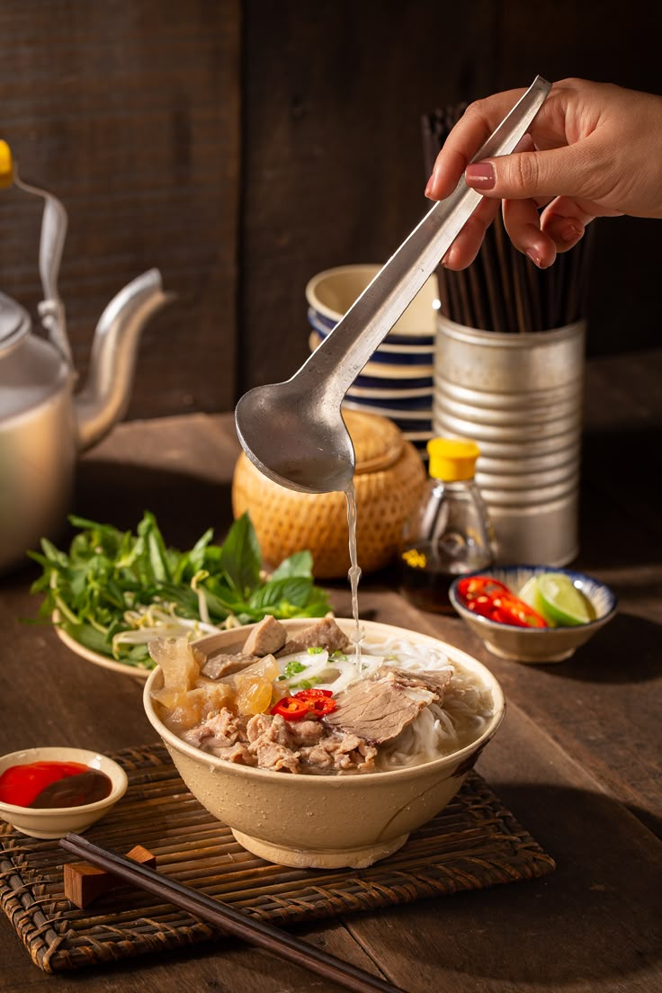 a person pouring soup into a bowl with chopsticks and sauce on the side