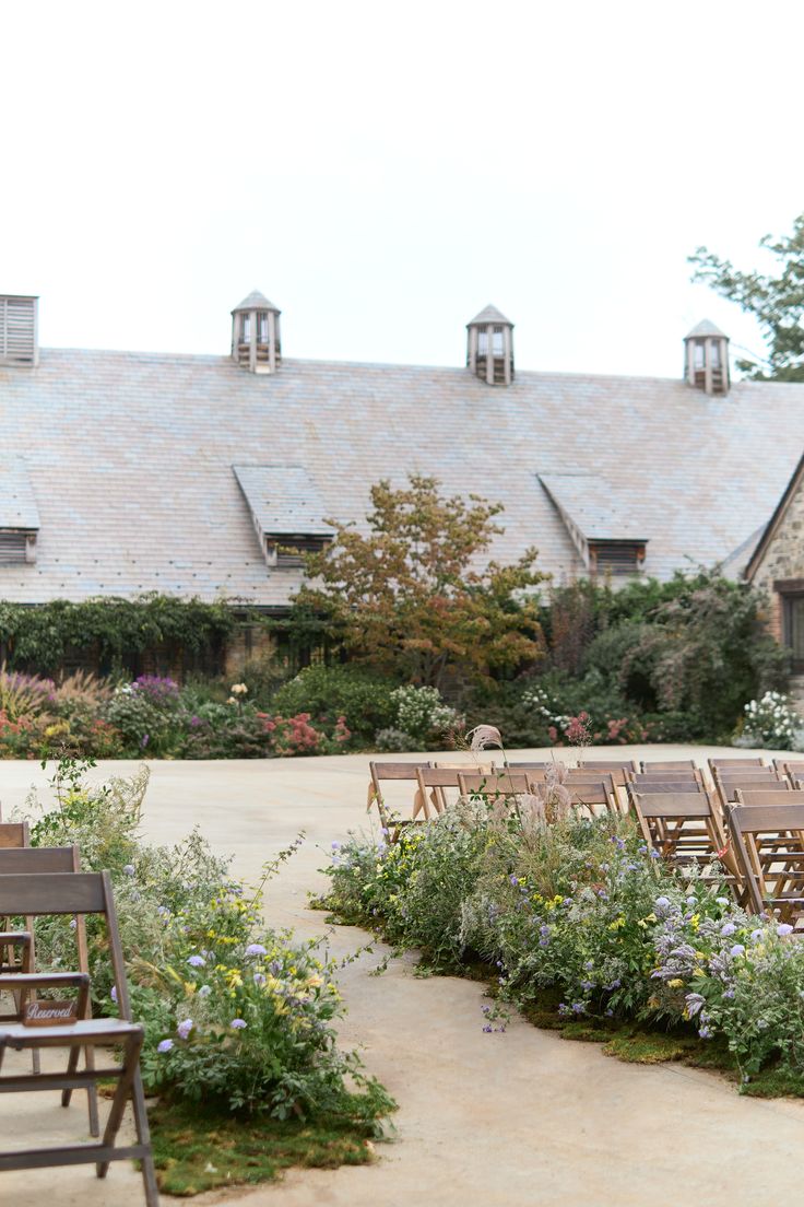 rows of wooden chairs are lined up in front of an old building with flowers and greenery