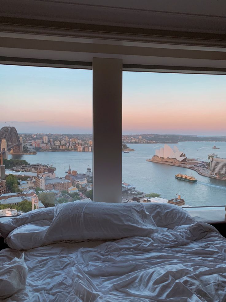 an unmade bed in front of two large windows overlooking the water and cityscape