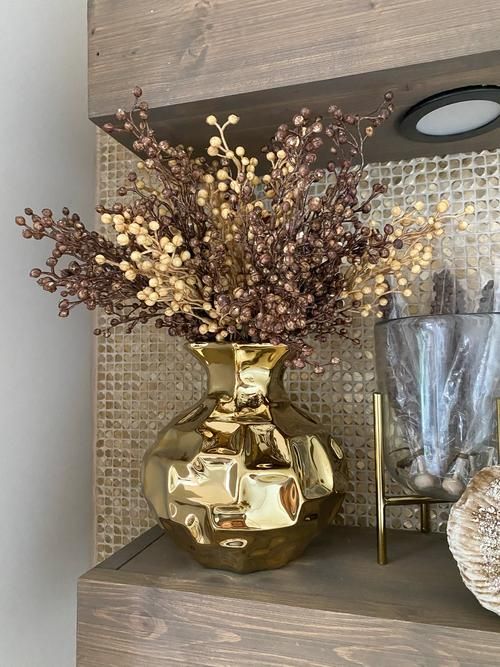 a gold vase filled with dried flowers on top of a shelf next to a lamp