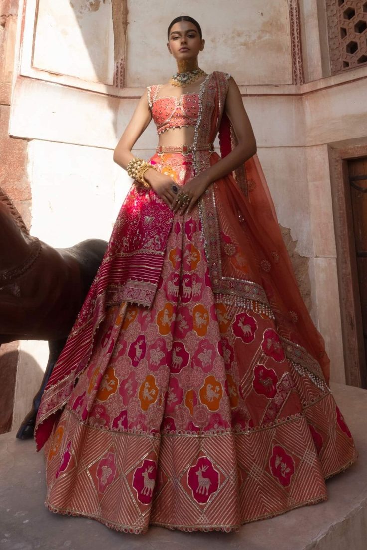 This stunning banarsi lehenga is sumptuously paired with a coral mirrored bralette-cut blouse, inspired by the enchanting Mughal gardens. Intricate blooming floral motifs and delicate fauna are beautifully scattered throughout, featuring charming butterflies and birds on the lehenga panel. The sides boast a playful texture of gota patti and motifs, adding a delightful twist to the design. Completing the look is the AG signature Meenakari belt at the waist, infusing the ensemble with elegance and sophistication. Banarasi Silk Sets With Gota Work For Reception, Reception Choli With Gota Work In Banarasi Silk, Banarasi Silk Choli With Gota Work For Reception, Reception Banarasi Silk Choli With Gota Work, Designer Meenakari Sharara In Chanderi, Pink Bollywood Pre-draped Saree With Meenakari, Art Silk Dupatta With Tilla For Reception, Reception Art Silk Dupatta With Tilla Detail, Designer Chanderi Lehenga With Tilla Embroidery