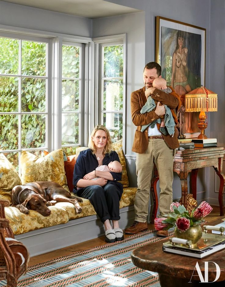 a man standing next to a woman holding a baby in her arms while sitting on a couch