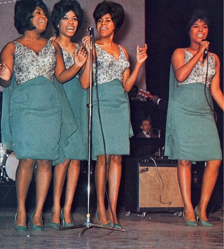 four women singing into a microphone while standing in front of a stage with other people