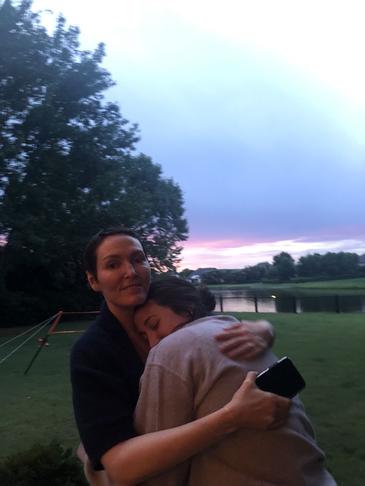 a woman holding a baby in her arms while standing next to a lake at dusk