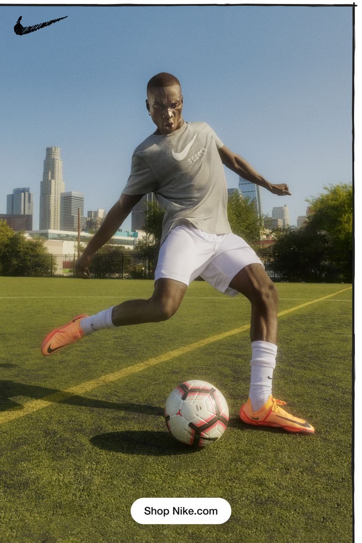 a man kicking a soccer ball on top of a green field with buildings in the background