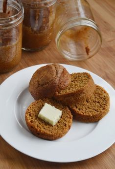 three muffins on a white plate with butter and jam in jars behind them