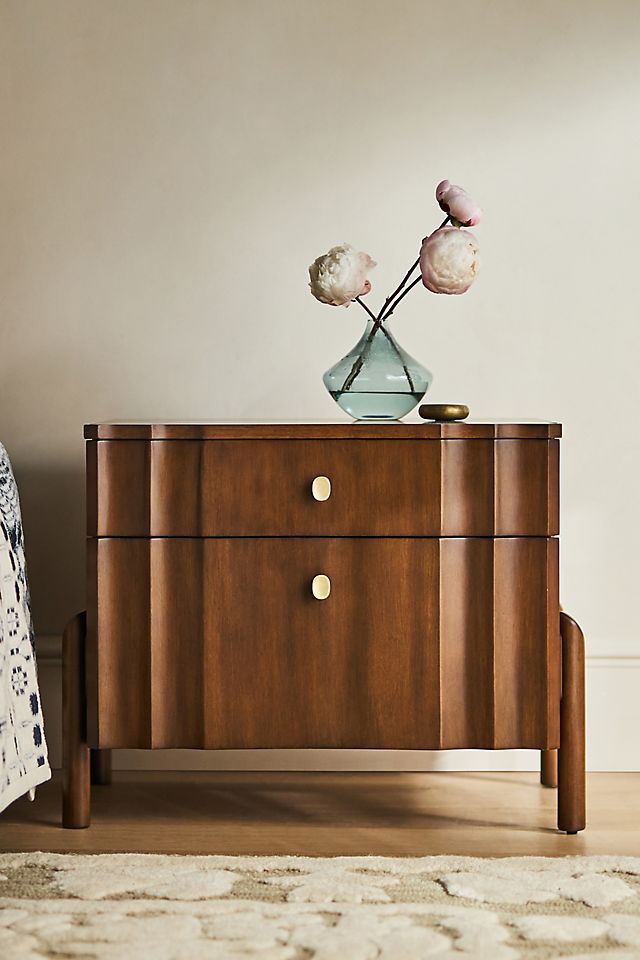 a wooden cabinet with two flowers on top and a bed in the back ground next to it