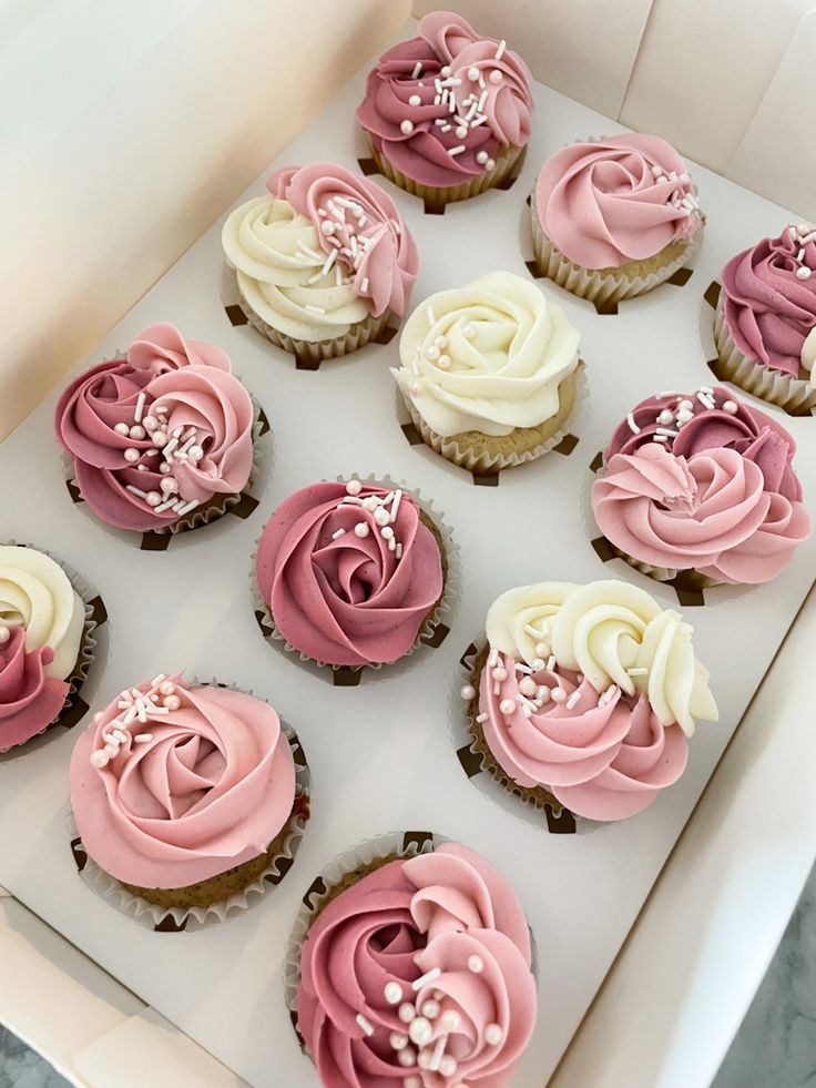 twelve cupcakes with pink and white frosting in a box on a table