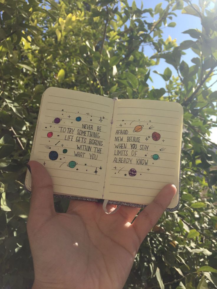 a hand holding an open notebook with writing on it in front of some trees and bushes