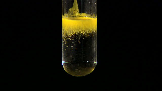 a yellow substance in a test tube filled with liquid and gold flakes on a black background