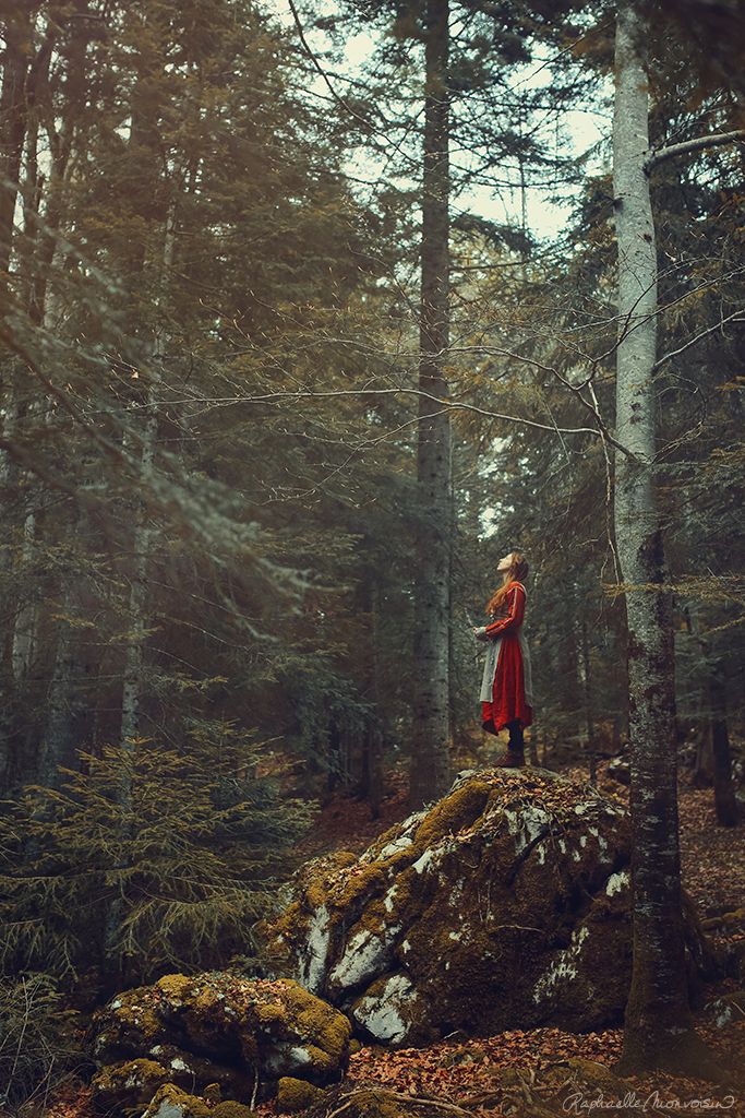 a woman standing on top of a rock in the woods with trees around her and wearing a red coat