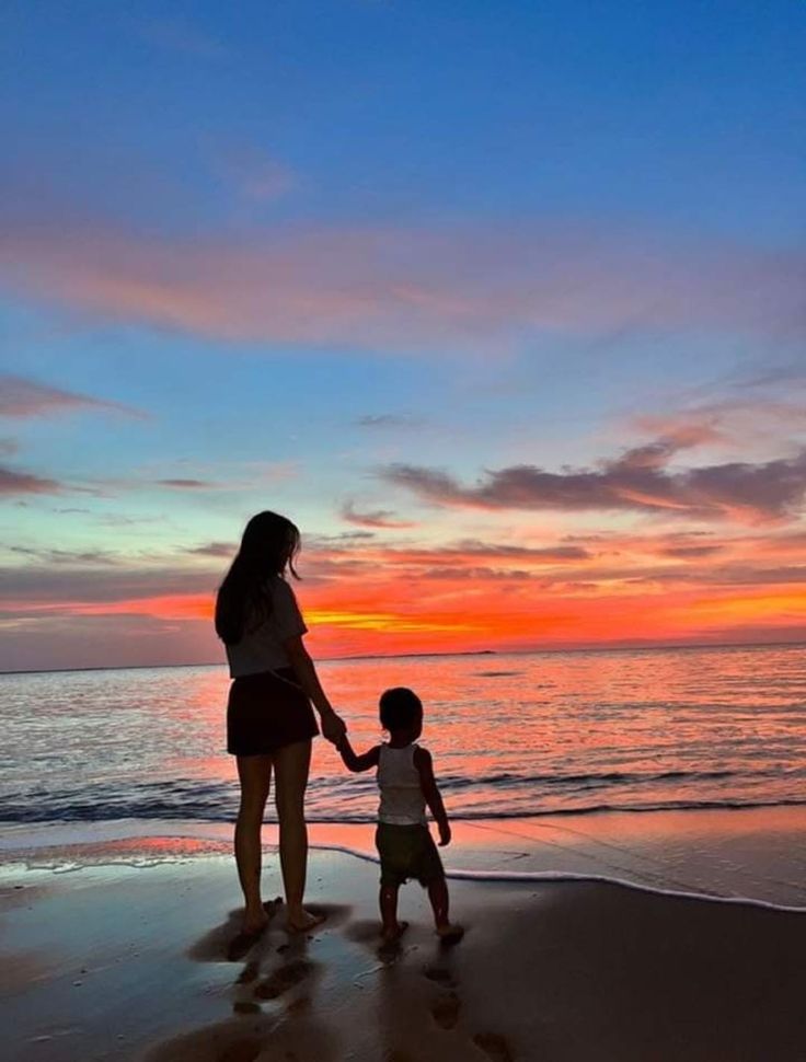 a woman holding the hand of a small child on top of a beach at sunset