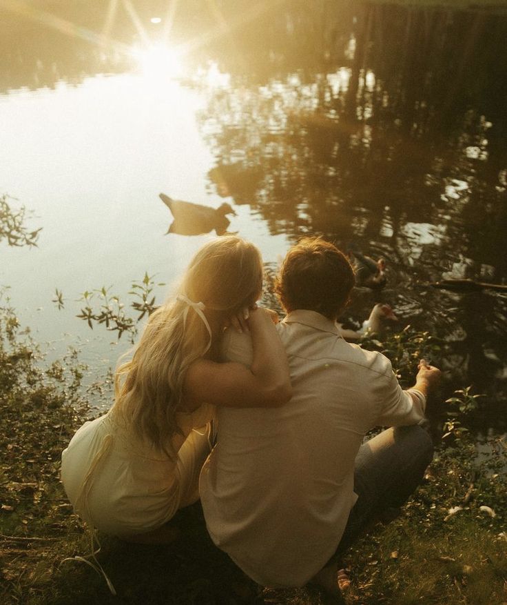 a man and woman sitting next to each other on the grass near water with ducks