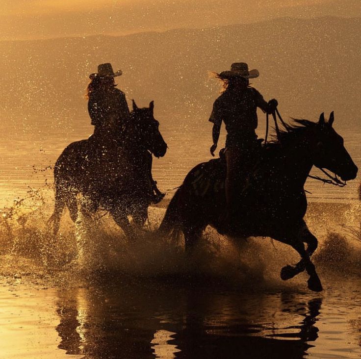 two people riding horses through the water at sunset