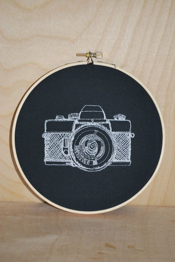 a black and white embroidered picture of a camera on a wooden surface with wood frame