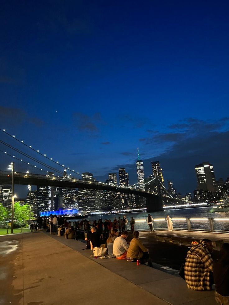 people are sitting on the edge of a walkway at night with city lights in the background