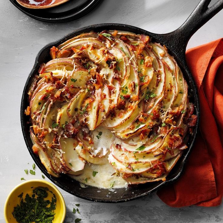 a skillet filled with potatoes and bacon on top of a white table next to bowls of sauces