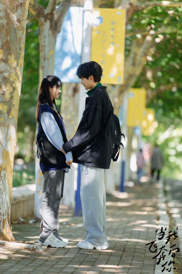 two people standing next to each other on a sidewalk with trees and signs in the background