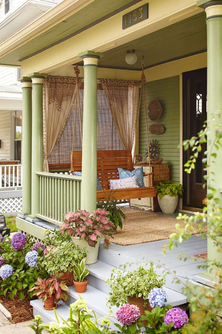 a porch covered in lots of plants and flowers next to a wooden bench with curtains on it