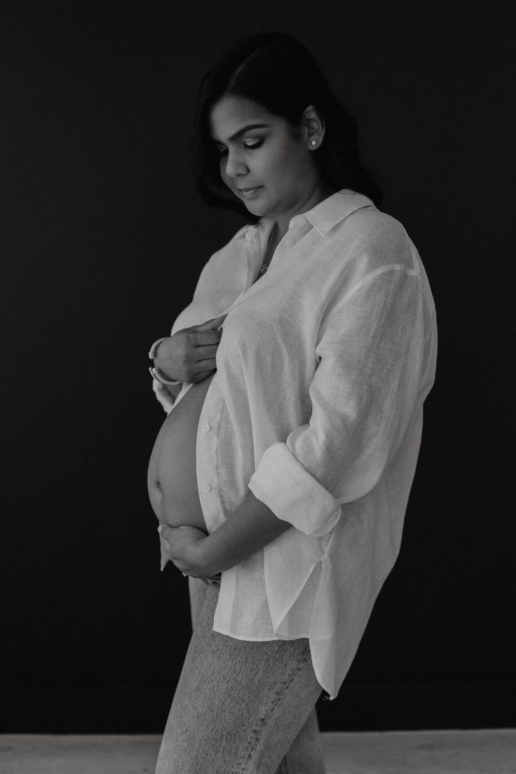 black and white photograph of a woman holding her pregnant baby bump while standing in front of a dark background