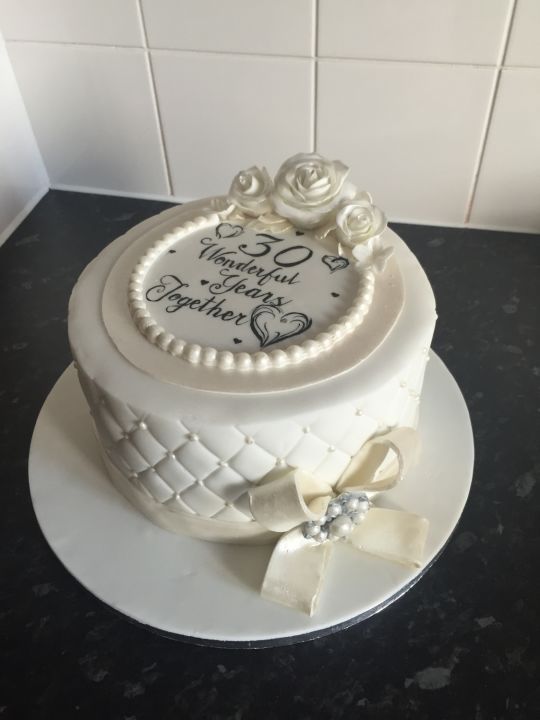 a white wedding cake decorated with roses and ribbons on a countertop next to a tile wall