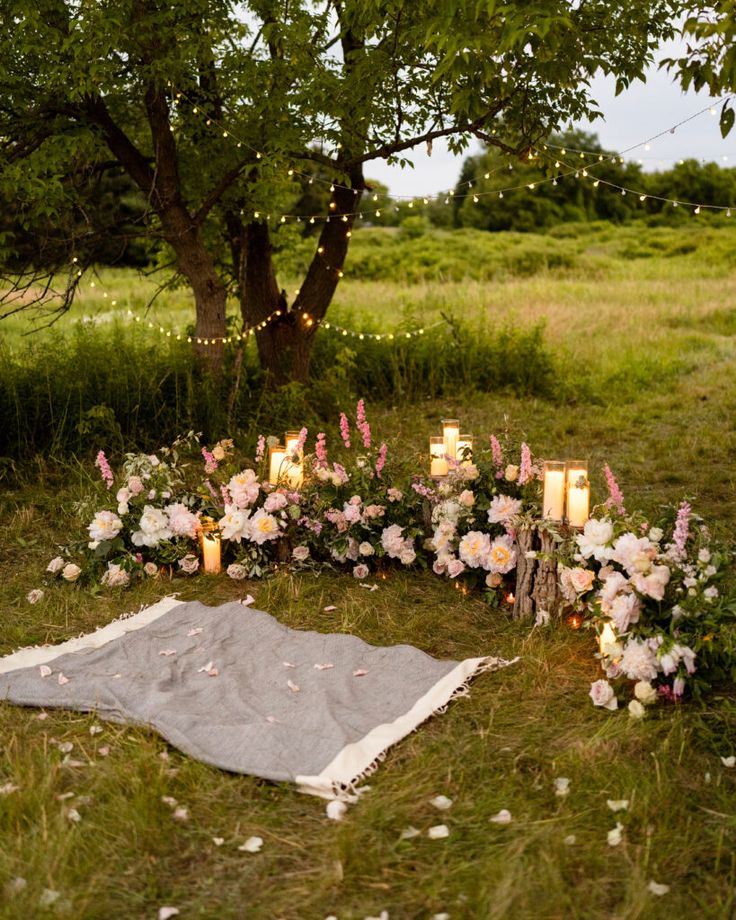 a blanket and flowers on the ground with candles in front of it, surrounded by greenery